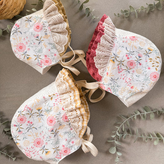Three floral baby bonnets on a neutral background with green leaves.