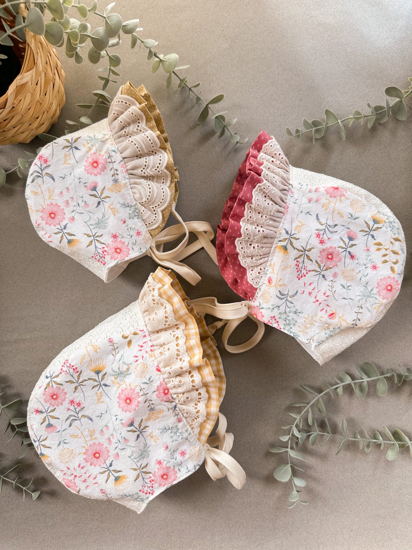 Three floral baby bonnets on a neutral background with greenery.