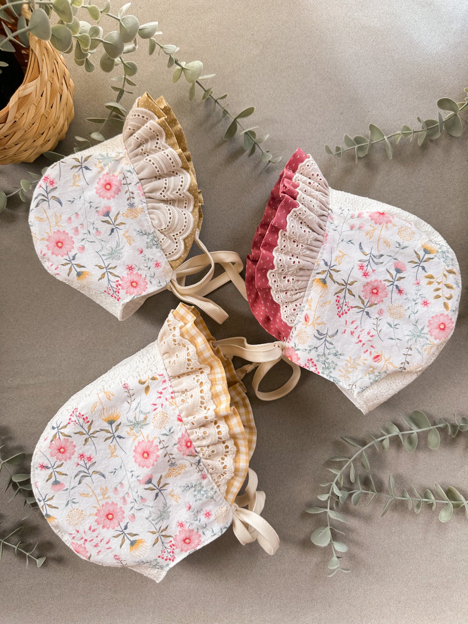 Three floral baby bonnets on a neutral background with greenery.