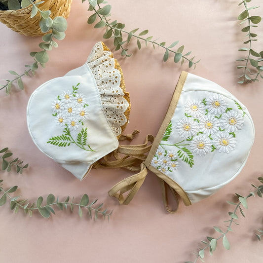 Two white baby bonnets with floral embroidery on a pink background with eucalyptus leaves.