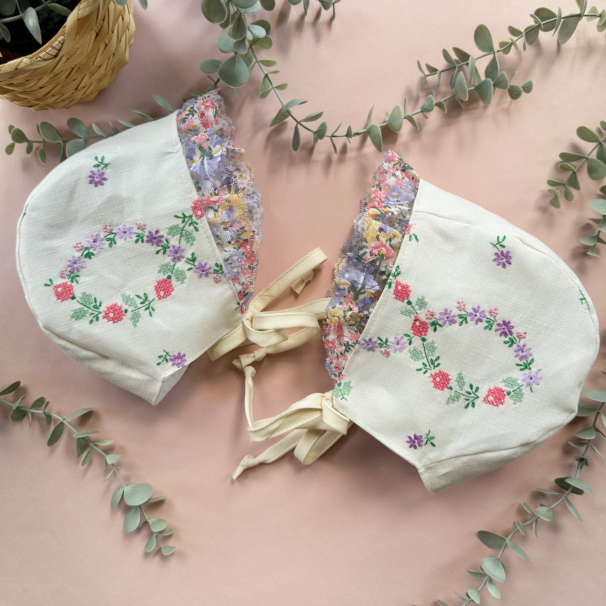Two floral embroidered fabric bonnets on a pink surface with green leaves.
