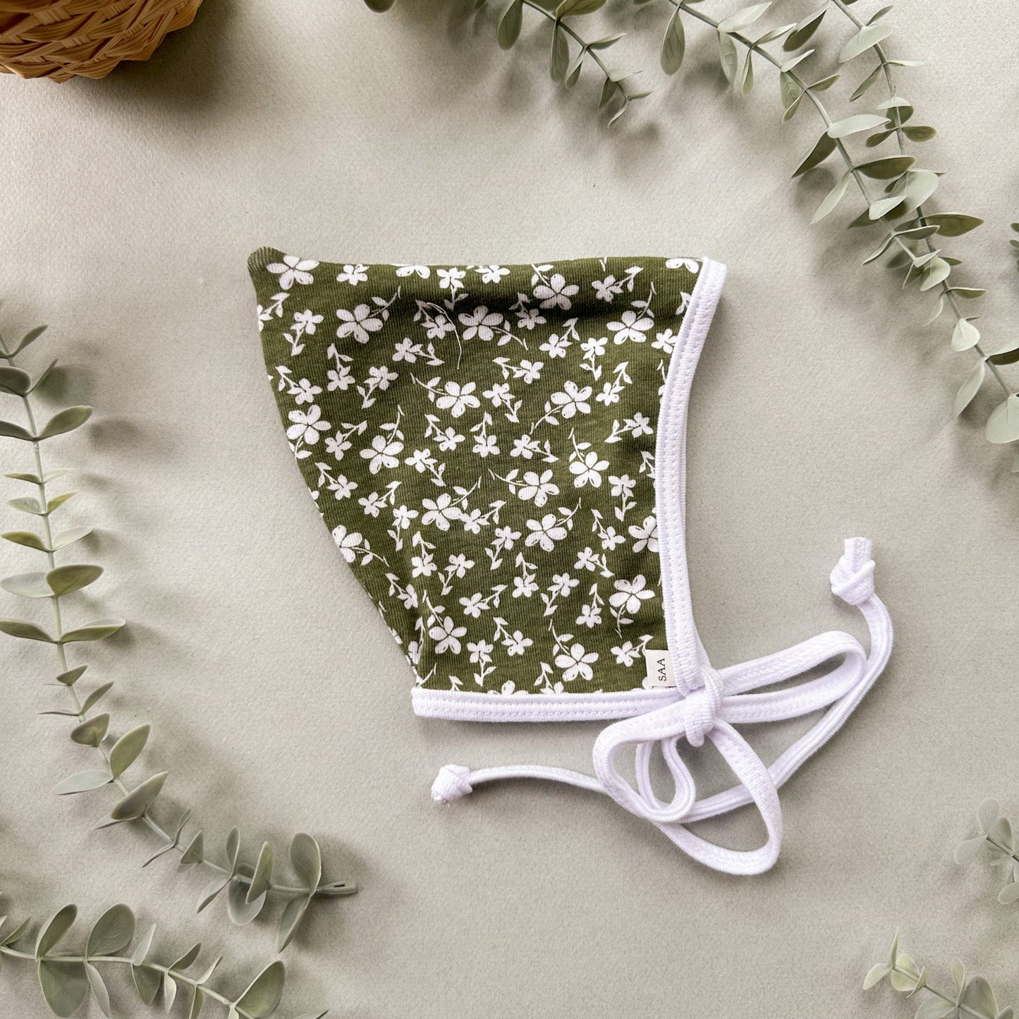Green floral baby bonnet with white ties on a light background with eucalyptus leaves.