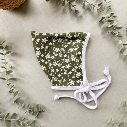Green floral baby bonnet with white ties on a light background with eucalyptus leaves.