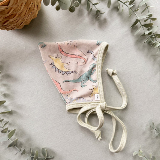 Baby bonnet with dinosaur design on a light gray background with eucalyptus leaves.