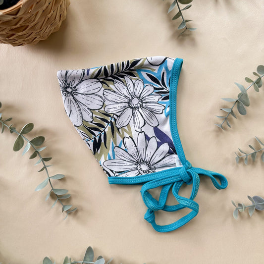 Floral bonnet with blue trim on a beige surface with eucalyptus leaves.