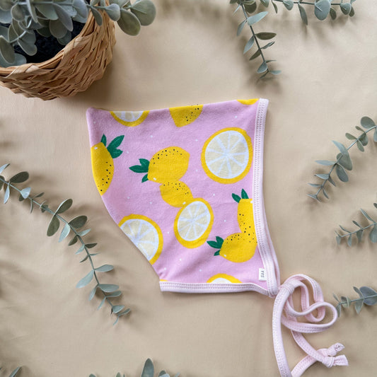 Pink baby bonnet with lemon pattern on a beige surface with eucalyptus leaves.