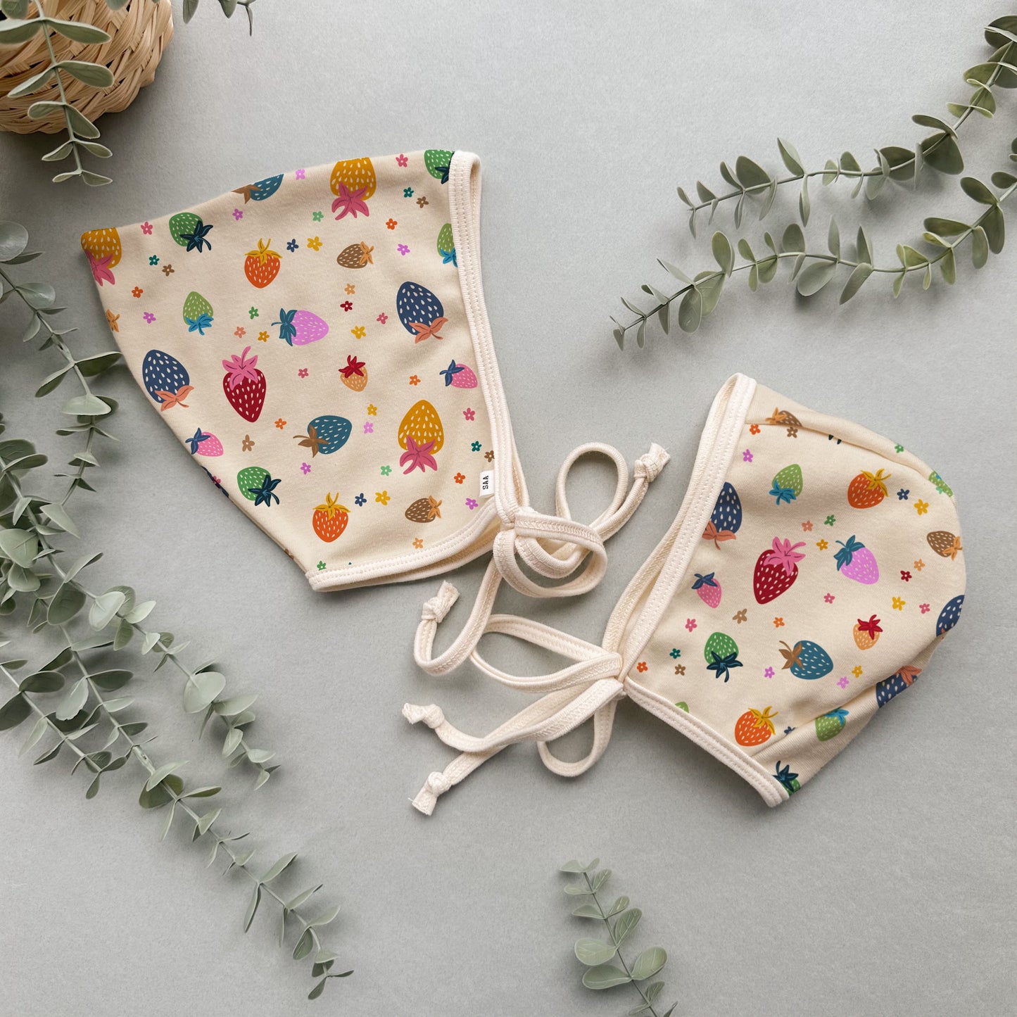 Two baby hats with colorful fruit pattern on a light gray background with green leaves.