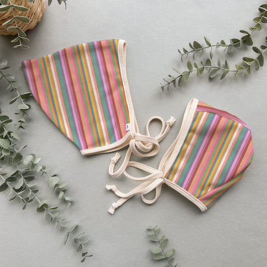 Pair of colorful striped organic cotton bonnets on a light gray background with green leaves.