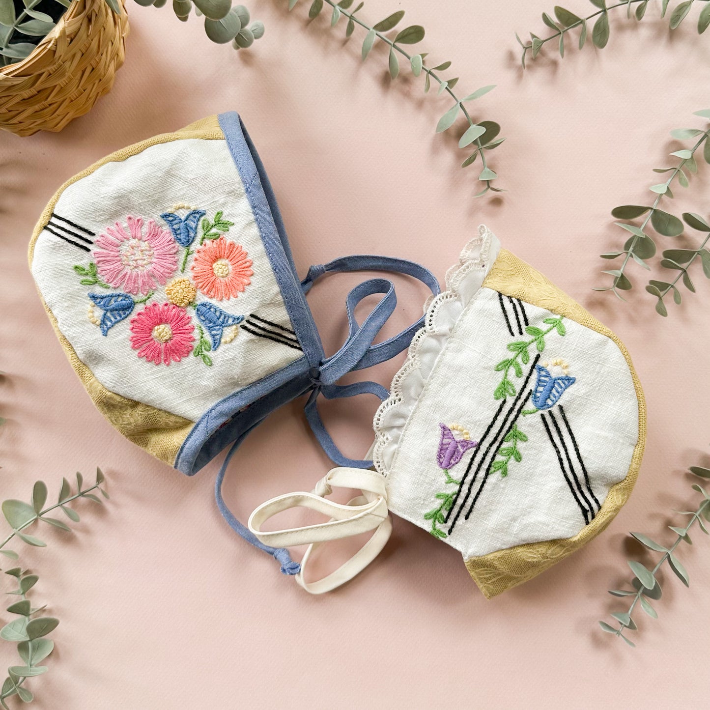 Two small embroidered fabric pouches or hats with blue ties, laid flat on a pink background with eucalyptus leaves and a woven basket around them. The fabric is off-white with floral and bird embroidery in pink, blue, green, yellow, and purple, plus black stripe details.