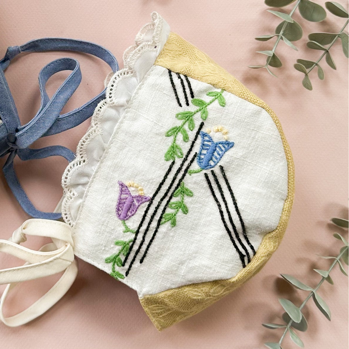 Two small embroidered fabric pouches or hats with blue ties, laid flat on a pink background with eucalyptus leaves and a woven basket around them. The fabric is off-white with floral and bird embroidery in pink, blue, green, yellow, and purple, plus black stripe details.