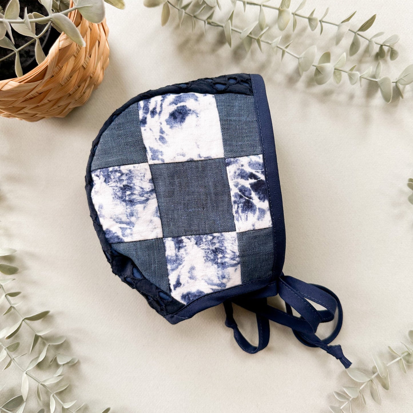 A handmade patchwork bonnet with a blue and white indigo tie-dye pattern, styled on a light beige surface with eucalyptus leaves and a small woven basket.