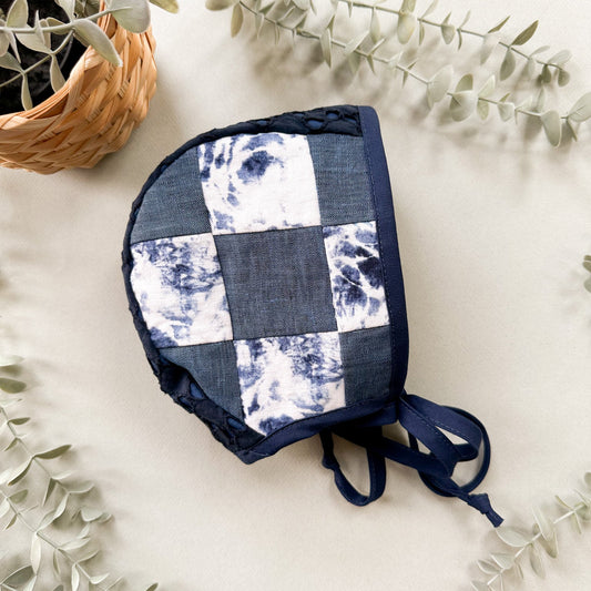A handmade patchwork bonnet with a blue and white indigo tie-dye pattern, styled on a light beige surface with eucalyptus leaves and a small woven basket.