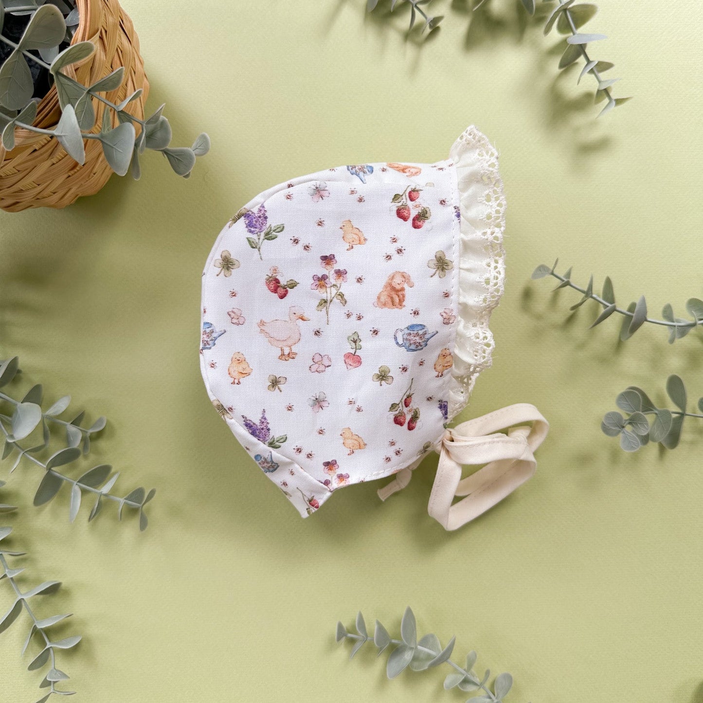 A baby bonnet with a white background and a colorful duck and floral print, styled on a light green background with eucalyptus leaves and a wicker basket.