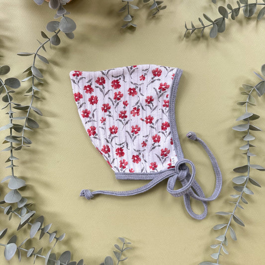 A baby bonnet with a white background, red floral print, and light gray knit trim and ties, styled flat-lay on a beige background with eucalyptus leaves.