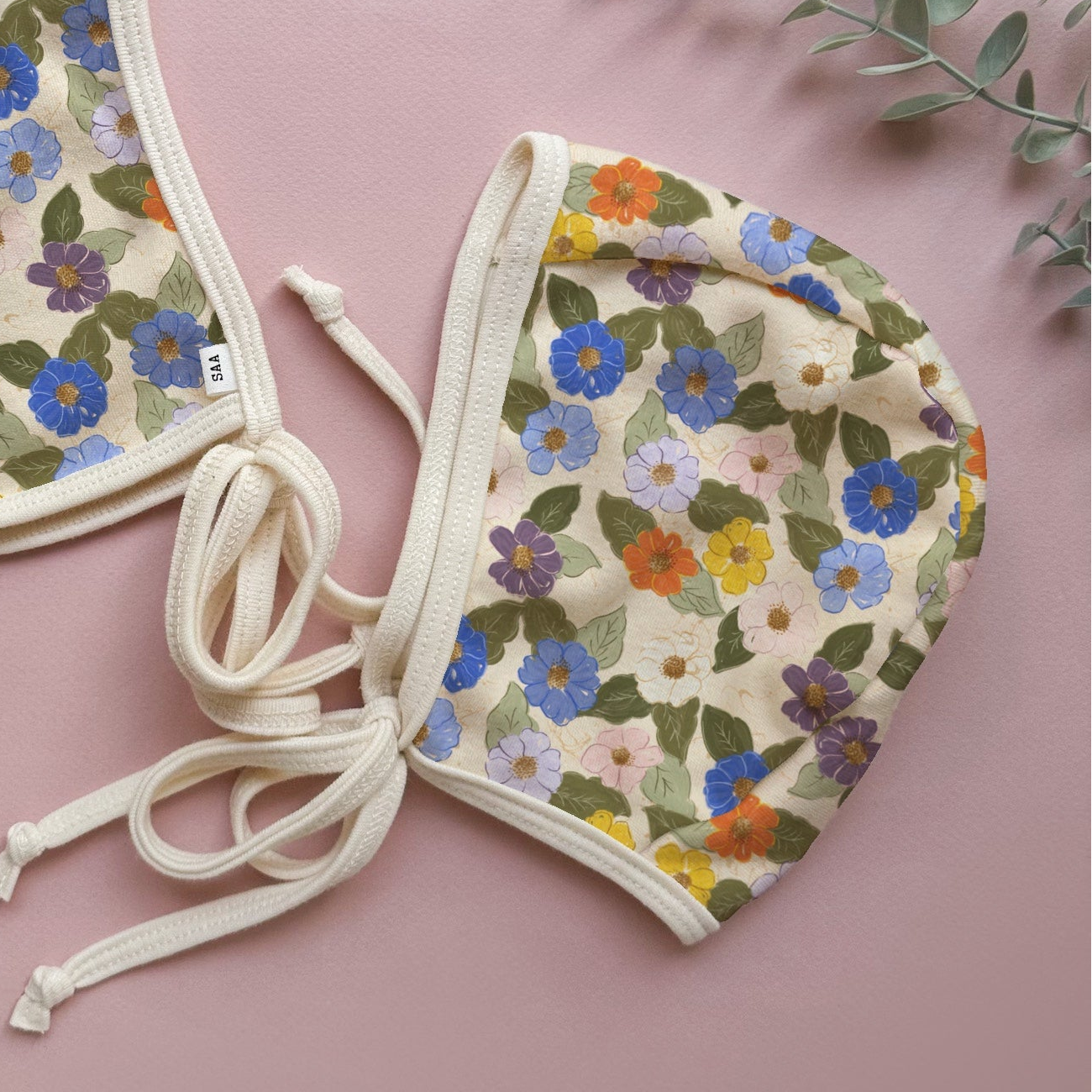 A floral-patterned knit bonnet displayed on a pale background, with a drawstring at the back. The bonnet is positioned towards the foreground, and there are decorative foliage accents in the background.