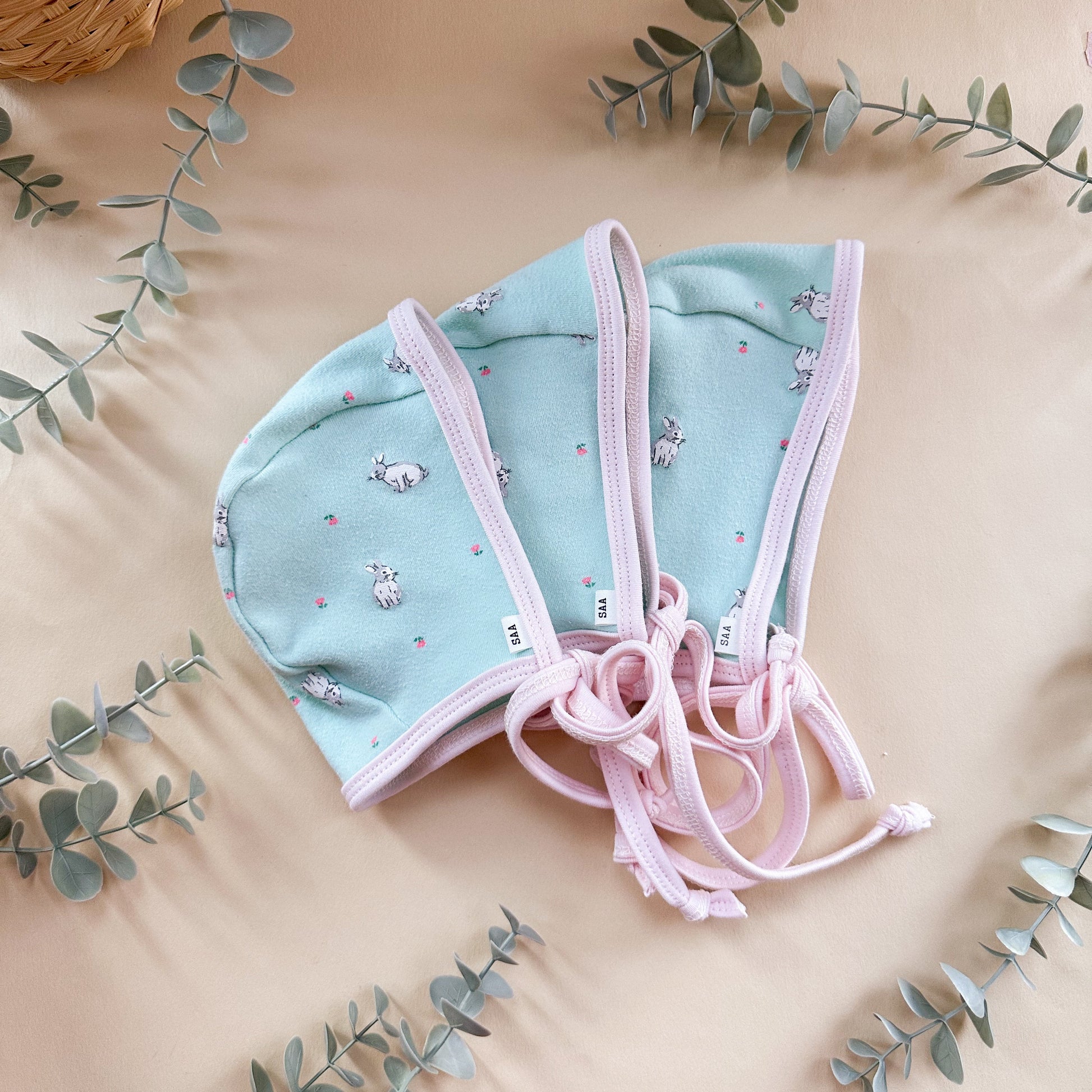 Light blue baby bonnet with pink trim on a beige background with eucalyptus leaves.