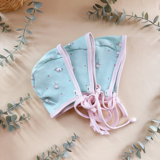 Light blue baby bonnet with pink trim on a beige background with eucalyptus leaves.