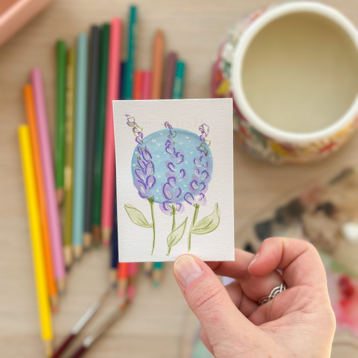 Hand holding a card with a blue flower drawing, surrounded by colored pencils and a mug. ACEO original artwork by Carrie Keefe - Bloom by April