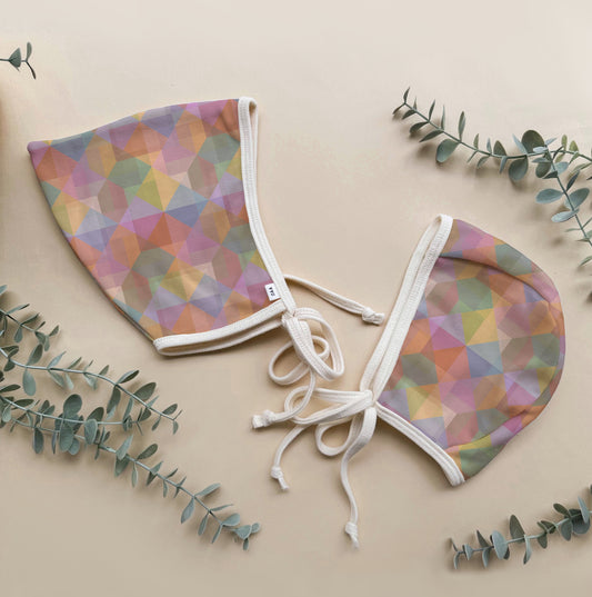 Two colorful baby bonnets with geometric patterns on a beige background with green leaves.