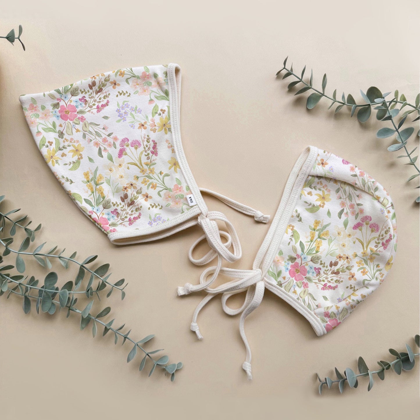 Floral baby bonnets set on a beige background with eucalyptus leaves.