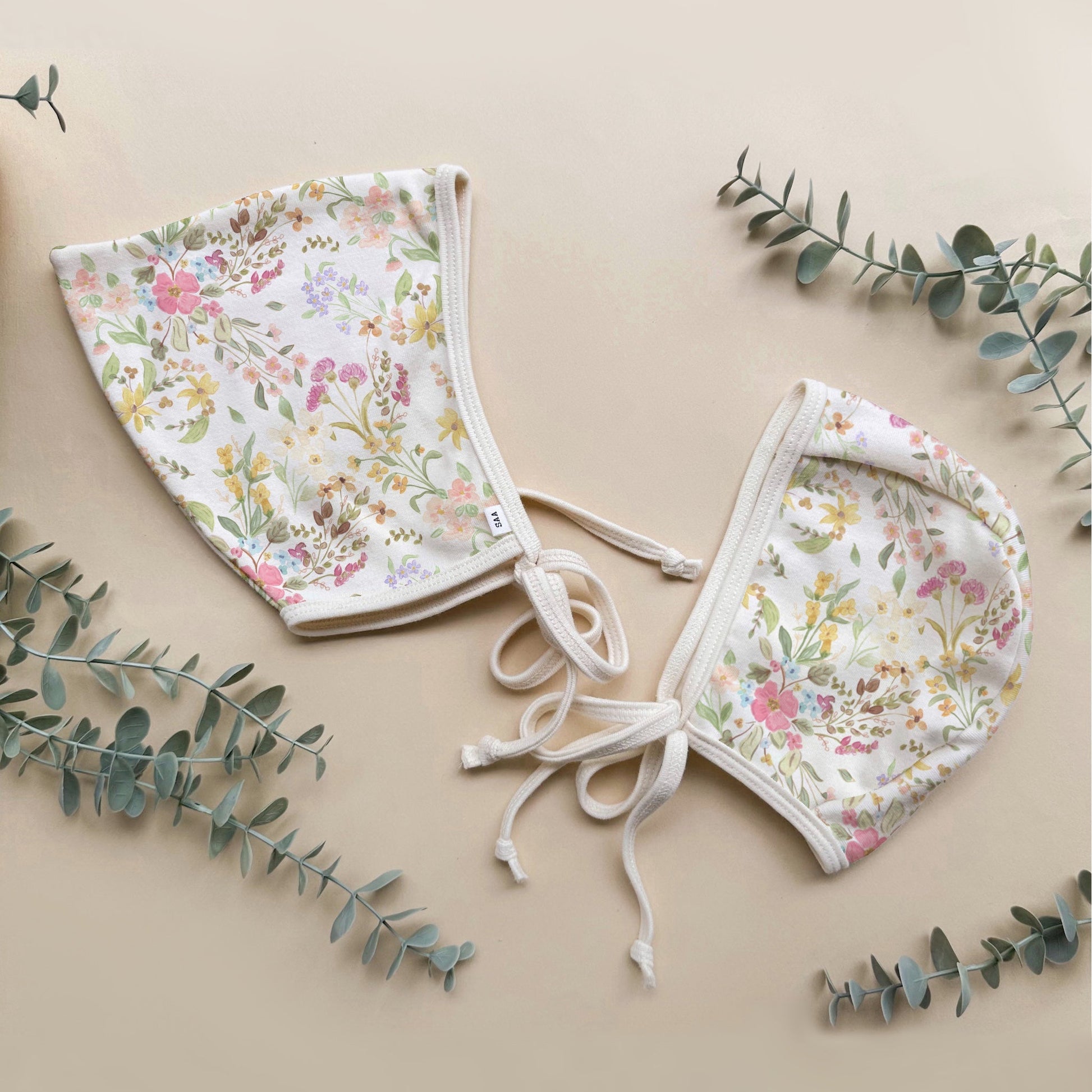 Floral baby bonnets set on a beige background with eucalyptus leaves.