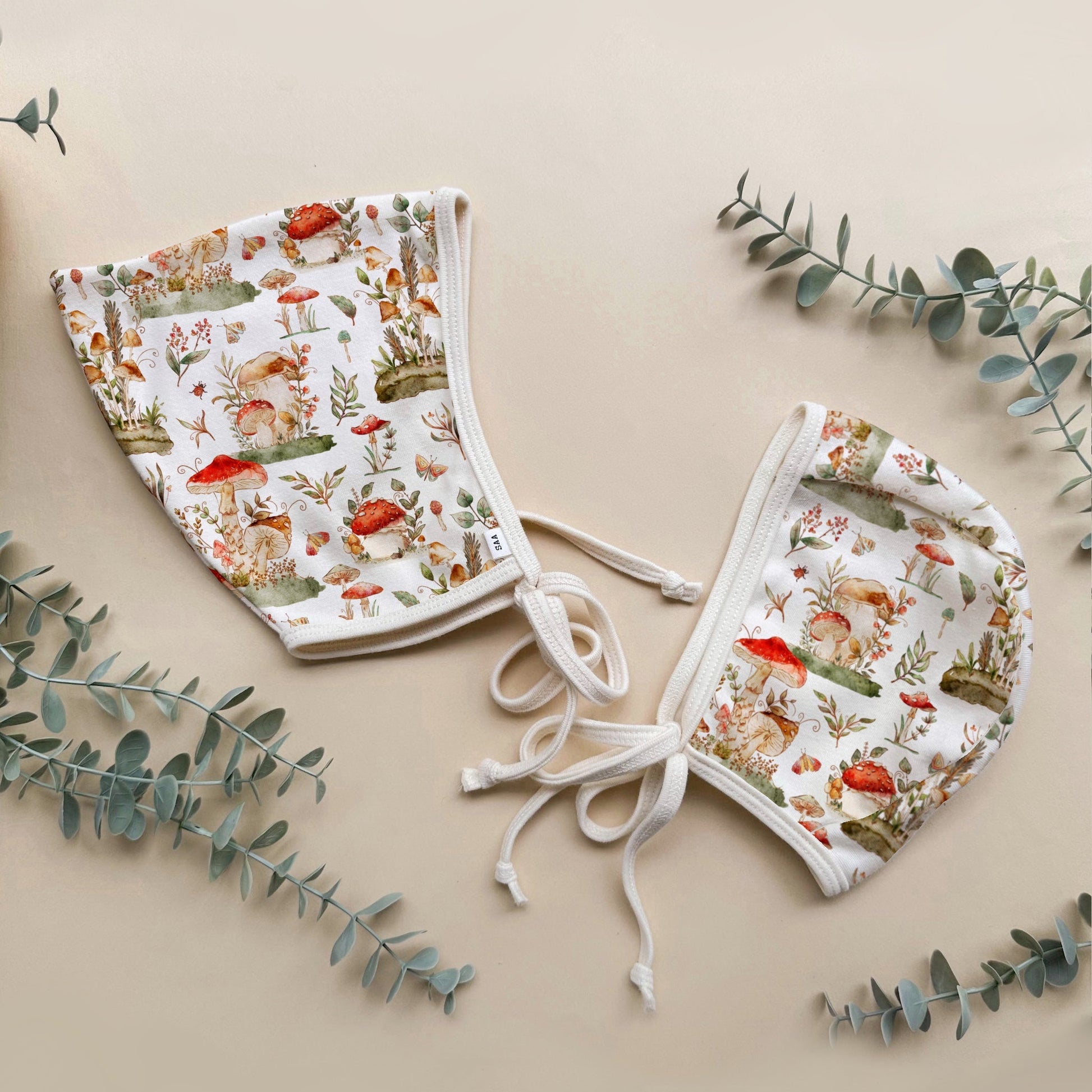 Two baby bonnets with mushroom nature-themed print on a beige background with eucalyptus leaves.