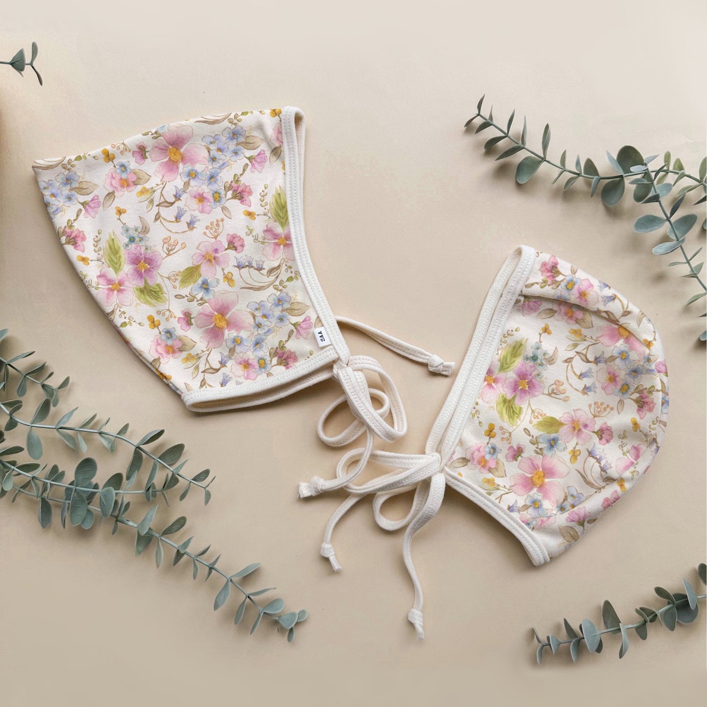 Two floral baby bonnets on a beige background with eucalyptus leaves.