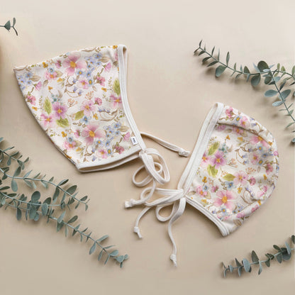 Two floral baby bonnets on a beige background with eucalyptus leaves.