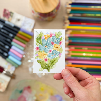 Hand holding a small card with a colorful floral design in front of colored pencils. ACEO original artwork by Carrie Keefe - Bloom by April