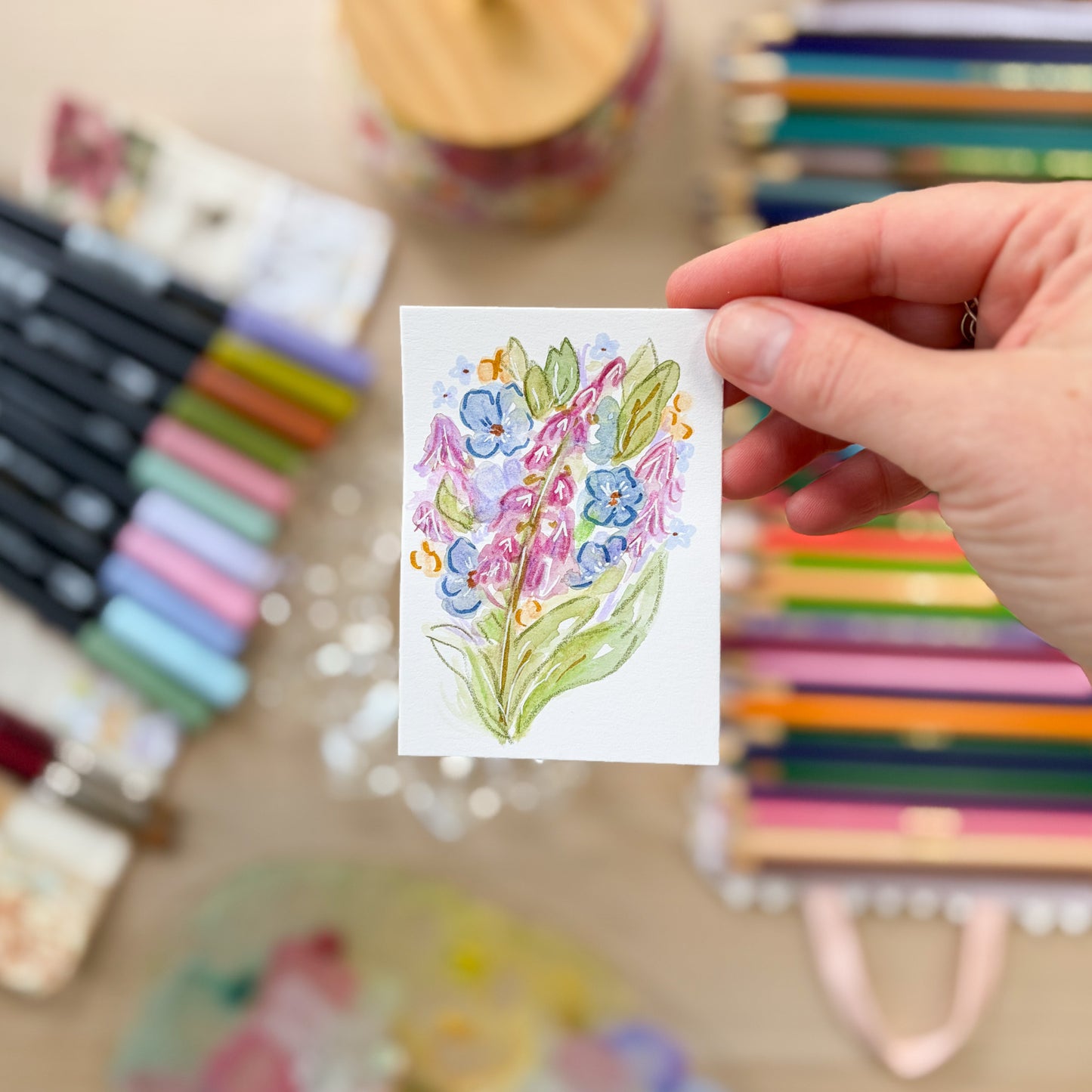 Hand holding a small watercolor painting of flowers with art supplies in the background. ACEO original artwork by Carrie Keefe - Bloom by April