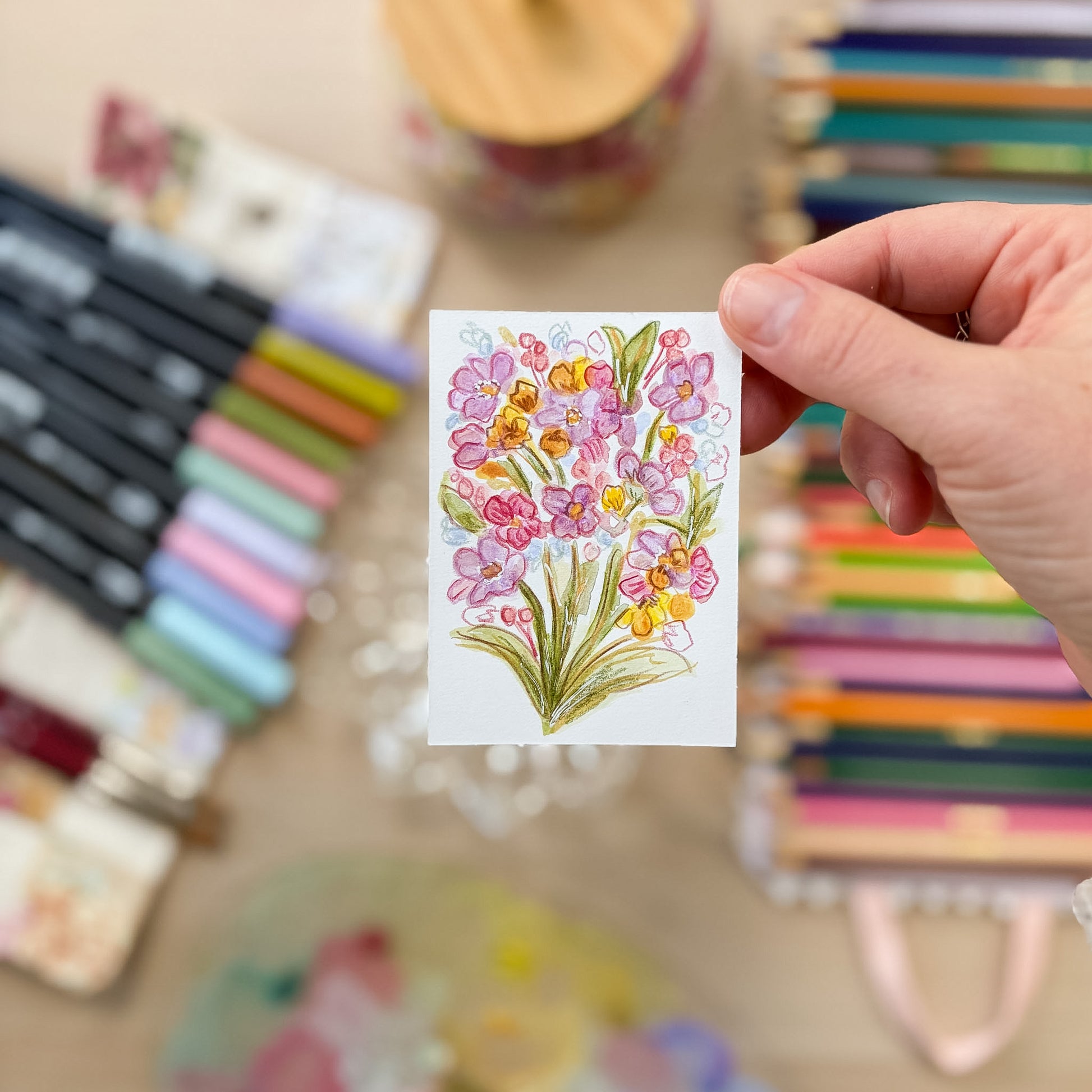 Hand holding a card with a floral design surrounded by colorful markers on a blurred background. ACEO original artwork by Carrie Keefe - Bloom by April