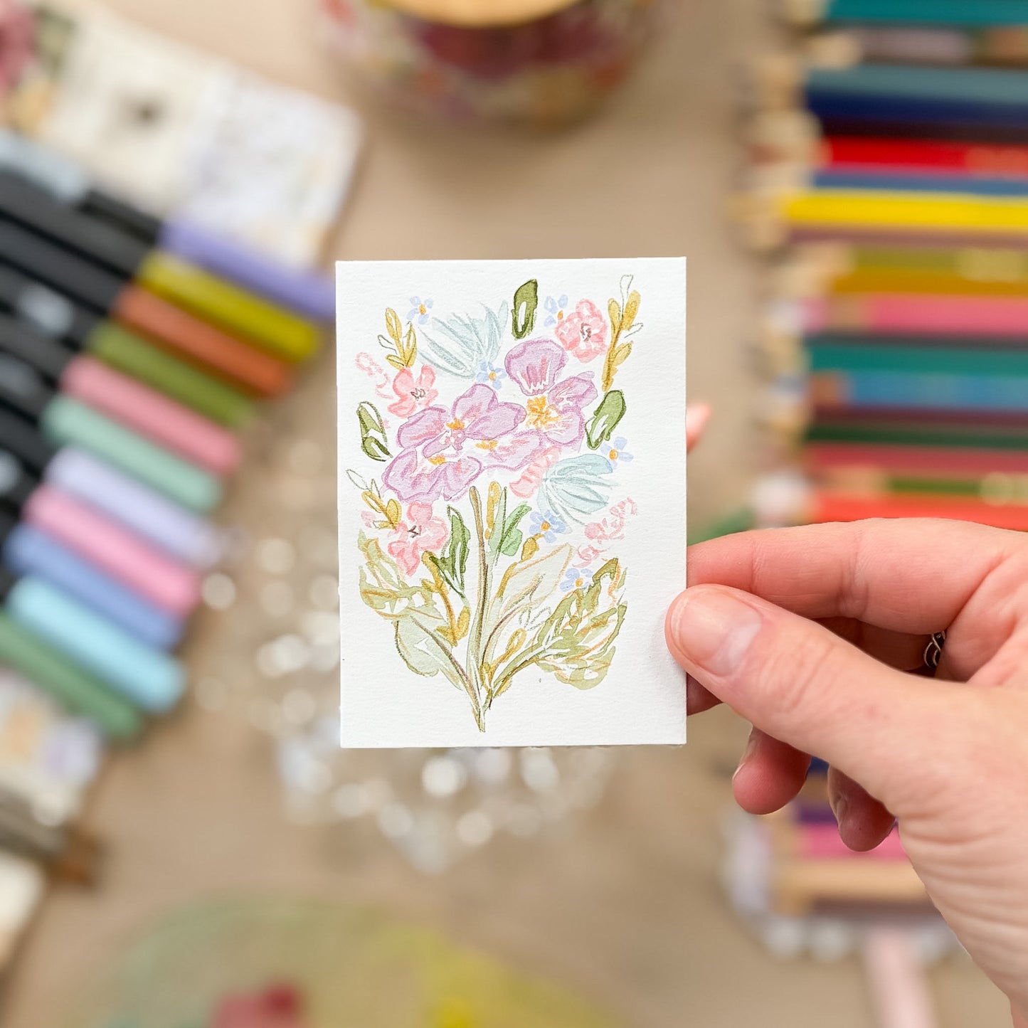 Hand holding a card with a floral design in front of colorful pencils. ACEO original artwork by Carrie Keefe - Bloom by April