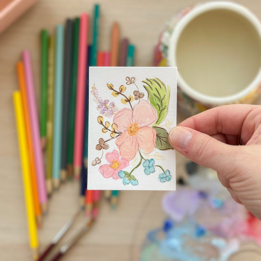 Hand holding a card with floral design surrounded by colored pencils and a mug. ACEO original artwork by Carrie Keefe - Bloom by April