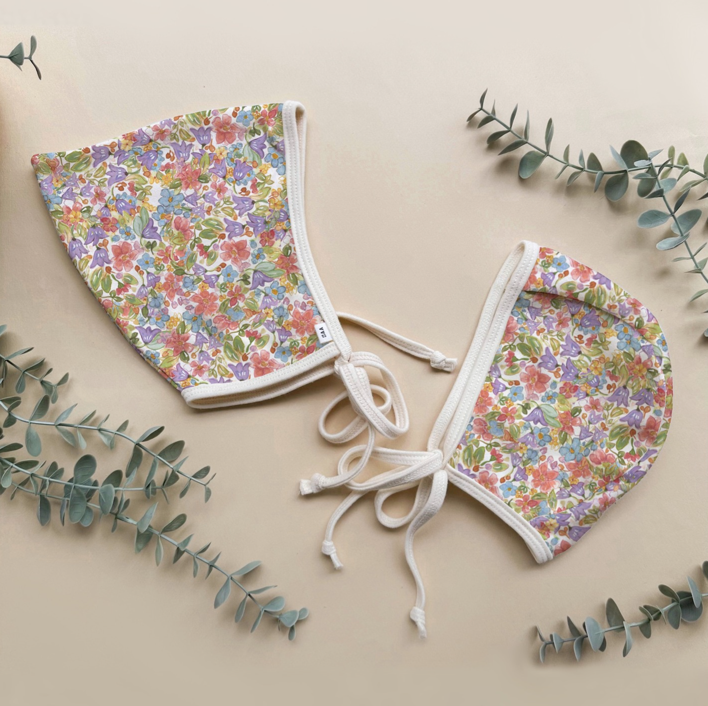 Floral-patterned baby bonnets on a beige background with green leaves.