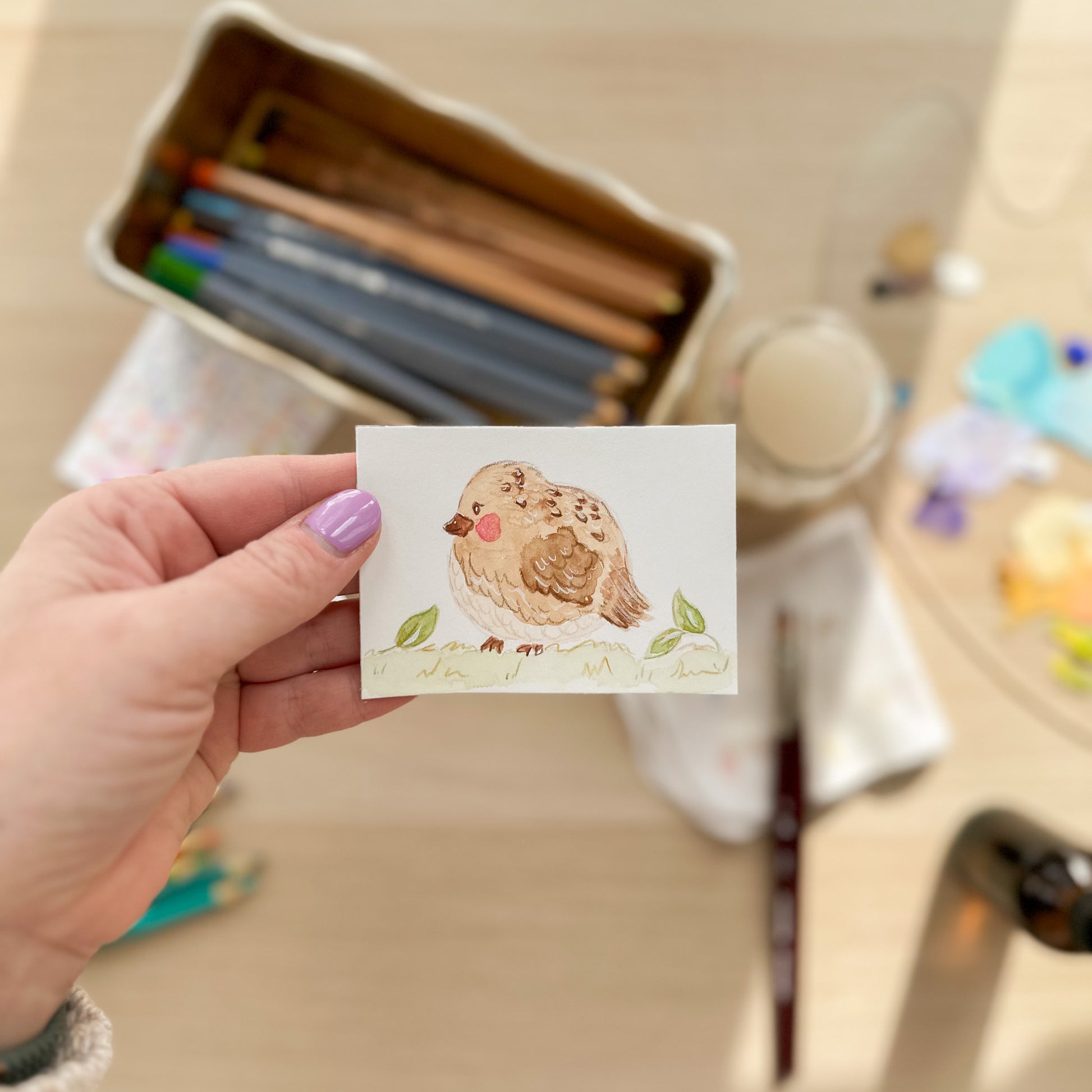 Hand holding a small watercolor painting of a bird with art supplies in the background. ACEO original artwork by Carrie Keefe - Bloom by April