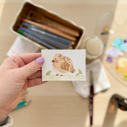 Hand holding a small watercolor painting of a bird with art supplies in the background. ACEO original artwork by Carrie Keefe - Bloom by April