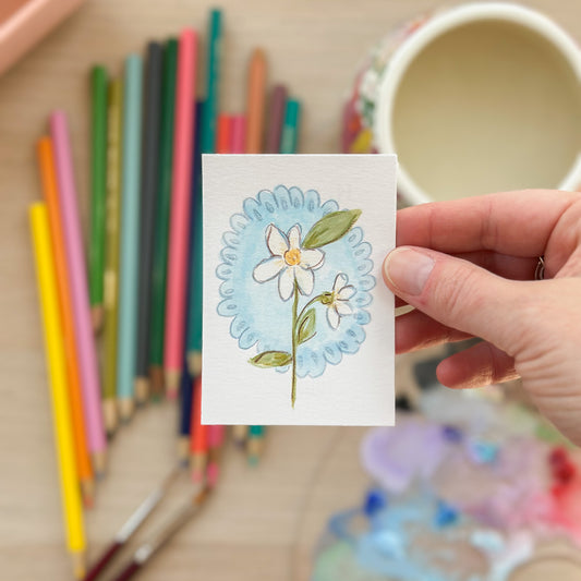 Hand holding a card with floral design surrounded by colorful pencils and a cup. ACEO original artwork by Carrie Keefe - Bloom by April