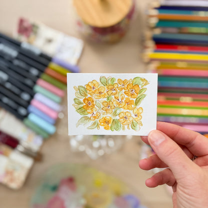 Hand holding a card with floral design surrounded by art supplies. ACEO original artwork by Carrie Keefe - Bloom by April