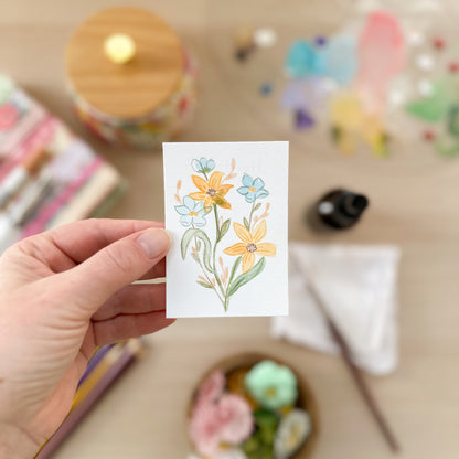 Hand holding a card with floral design on a blurred craft background. ACEO original artwork by Carrie Keefe - Bloom by April